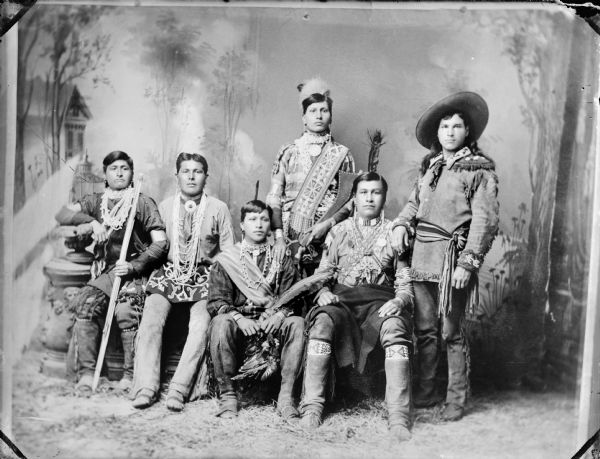 Studio portrait of six young Native American men, two standing and four sitting in native dress.