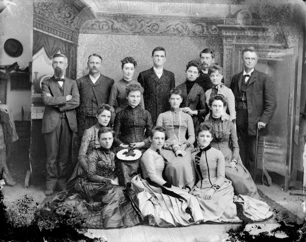 Studio portrait in front of a painted backdrop of five men posed standing, and ten women, three posed standing, three sitting, and four kneeling. Two standing men on the extreme left, from the left, probably Jones and G.M. Perry. Standing man on the far left is probably Dr. H.B. Cole.	