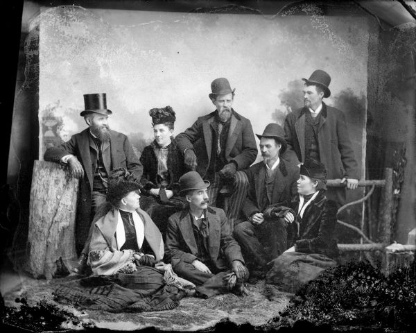Studio portrait in front of a painted backdrop of five men and three women posed next to a prop wooden fence.	