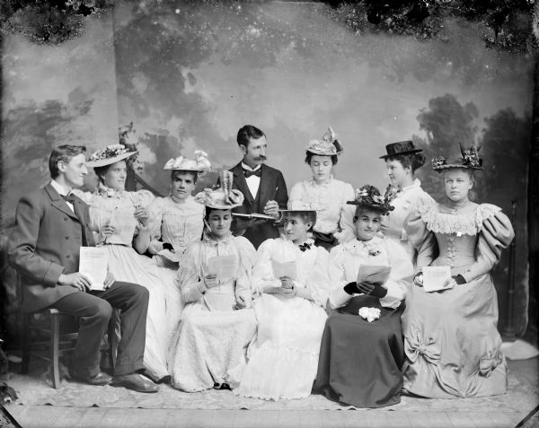 Studio portrait in front of a painted backdrop of two men and eight women posed sitting with books/magazines, The Intermediate Quarterly, possibly a discussion group. Woman sitting on a chair on the extreme right, and showing the title of the printed work, is probably Sadie Dimmick. Man and the two women sitting on the chairs to the extreme left is probably Haas Johnson and the Walters sisters.