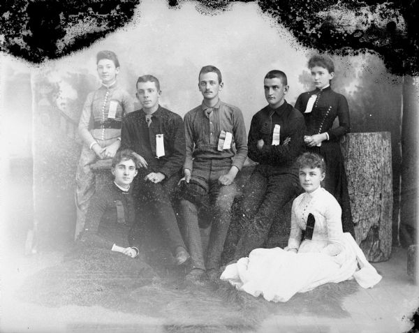 Studio portrait in front of a painted backdrop of three men and two women sitting in the center, and two women standing beside them on the left and right. The group has ribbons or badges pinned to the front of their clothing. The woman standing on the extreme right is probably Mrs. Merlin (Jessie) Hill.	