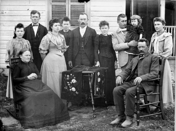 Elderly man and woman posed sitting on either side of a table in front of a wooden house. The couple is surrounded by a group of adults and one child. Three of them are men, one of whom is holding a young girl in his arms, the other five are women.