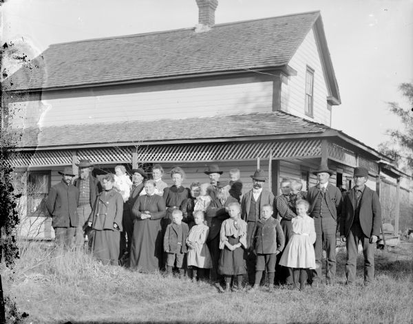 Group of men, women, and children posed standing along the side of a house. Second man standing from the left is probably Isaiah Landers, the third standing man from the left and holding the small child is probably Lou Darwin. Small boy standing in the front on the right is probably Ed Darwin.	