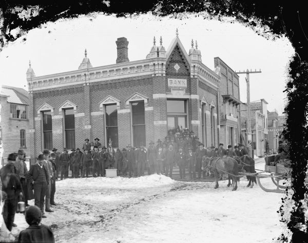 Large group of men and women, including a number of Native Americans, posed standing in front of the Jackson County Bank on a snow-covered street, board sidewalk and building steps, and a sled pulled by a team of two horses.
