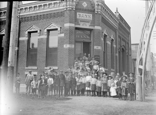 Large group of children posed standing in front of the Jackson Country Bank on the board sidewalk and building steps. Most are wearing school bags that the bank probably gave them. The man with the moustache in the doorway is probably Sam Lund, the director of the bank. A section of the arch in the intersection is on the right.