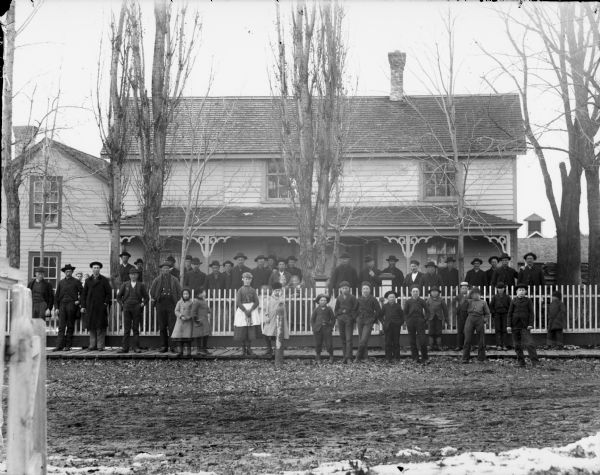Large group of men, and a few women and children, posed standing in front of a wooden building on both sides of a picket fence, probably the Prestemon Boarding House. Seventh boy in the front from the right is possibly Oscar Halvorson.	