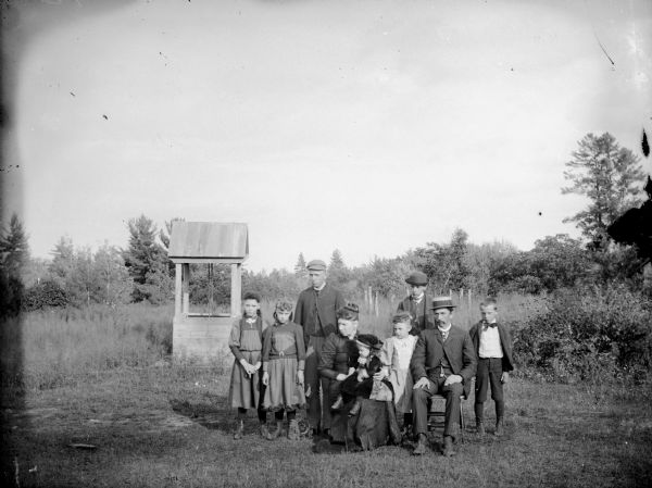 Group portrait of a man, and woman holding a small girl, posing sitting. They are surrounded by three girls and three boys standing near a well.
