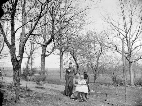 Group portrait of an elderly woman sitting and holding a small girl, in front of a young woman and girl standing just behind her. The group is posing among trees in a field.