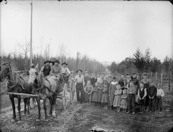 Large group of men, women and children posing standing next to five men posing sitting in a wagon pulled by a team of two horses on a country road. One boy appears to be using crutches.