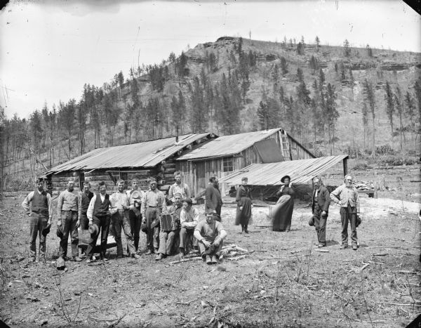 Two women and fourteen men posed standing and sitting in front of a log building, probably at the foot of "Silver Mound" near Hixton. One seated man is posed playing an accordion.