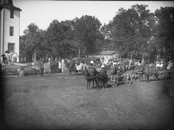 Slightly elevated view of a large group of people, some posing sitting in wagons on the road, and others standing near the fence, in front of a church, probably the Little Norway Church.