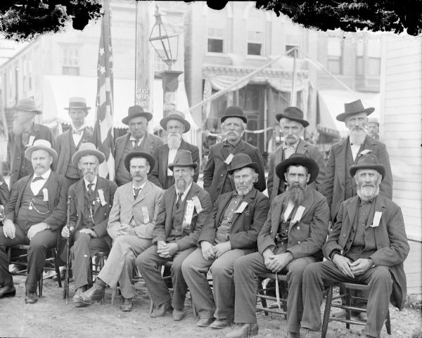 Elderly men posed sitting and standing on a street corner in town, probably a reunion of Civil War veterans.