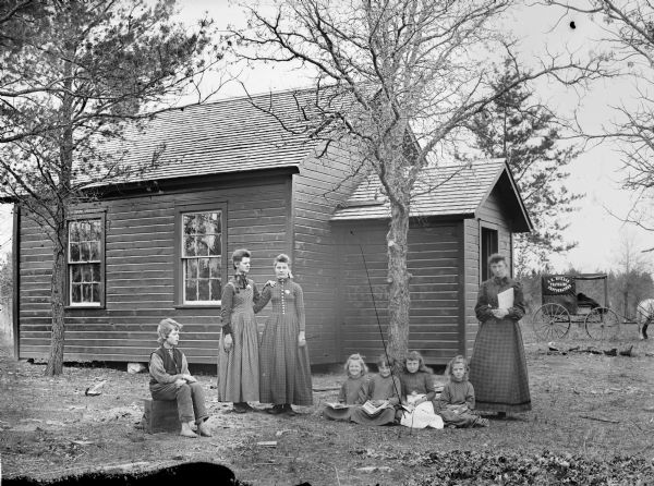 Group portrait of a female schoolteacher and two young women posing standing, and four girls and a boy posing sitting in front of a small schoolhouse, probably the Halcyon School. The photographer's wagon of C.R. Monroe is in the background.