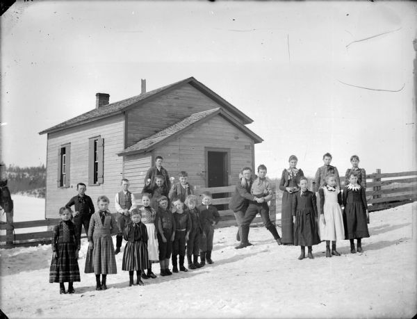 View towards a group of people, including a two boys grappling in the center, in front of a wooden building. Some of the people are holding apples. There is a wooden fence along the snow-covered road. Probably a school group.