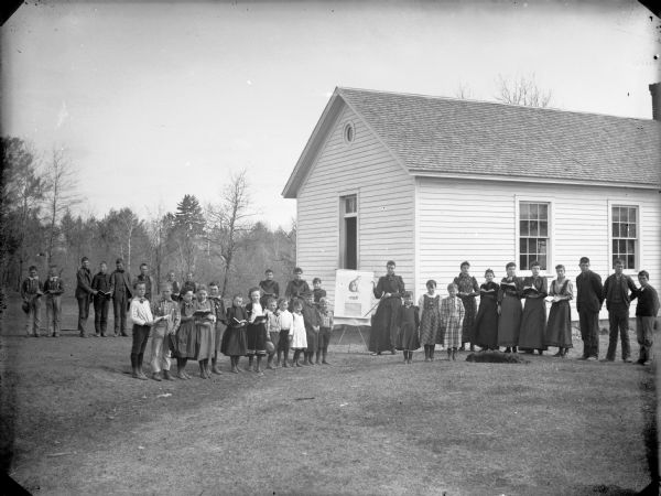 A teacher and students posing outdoors in the schoolyard of Shamrock School. The teacher is holding a reading chart and the children are holding books.