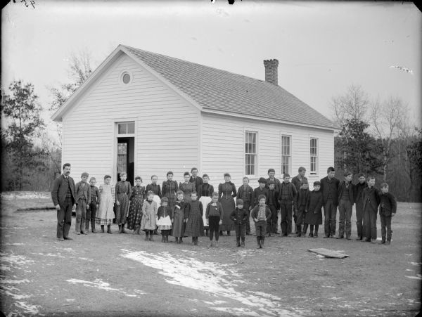 Male schoolteacher and a large group of boys and girls posing standing in front of a wooden building, probably a school group in front of the Shamrock School. The teacher is probably Herbert Perry.
