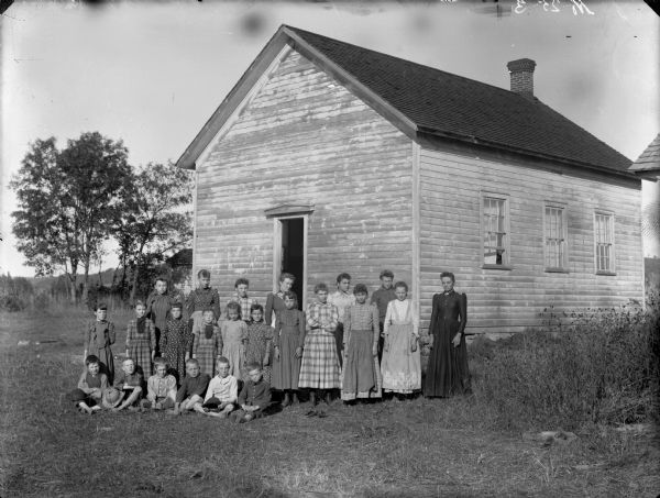 Group portrait of woman and a group of boys and girls posing sitting and standing in front of a wooden building, probably a school group. Negative has ink inscription, "No. 25 - 3."