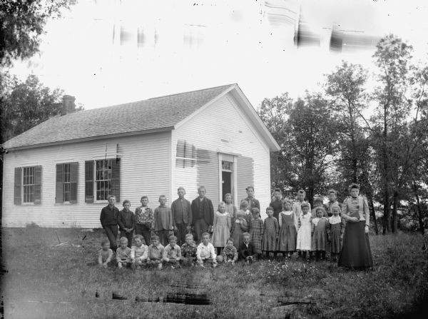 Group portrait of a woman and a group of boys and girls posing sitting and standing in front of a wooden building, probably a school group. Negative has ink inscription, "No. 4."	