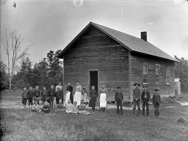 Boys and girls are posing sitting and standing in front of a wooden building, probably a school group in front of the Pole Grove School near Alma Center.