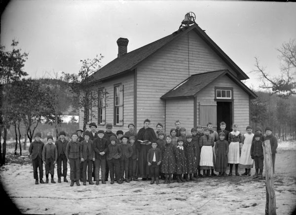 Group portrait of a woman and a group of boys and girls posing standing on the snow-covered ground behind a barbwire fence, in front of a wooden building with a bell on the roof, probably a school group.