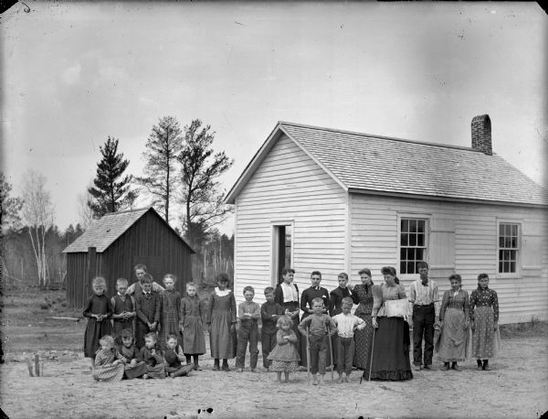Group portrait of a woman holding a broom and a book standing with a group of boys and girls. They are standing in front of a wooden building, probably a school group, possibly the Thomas School.	