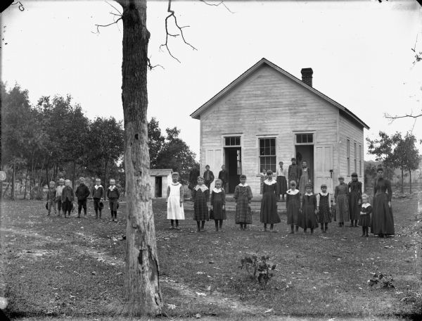Group portrait of a woman and a group of boys and girls posing standing behind a tree in front of a wooden building, probably a school group.	