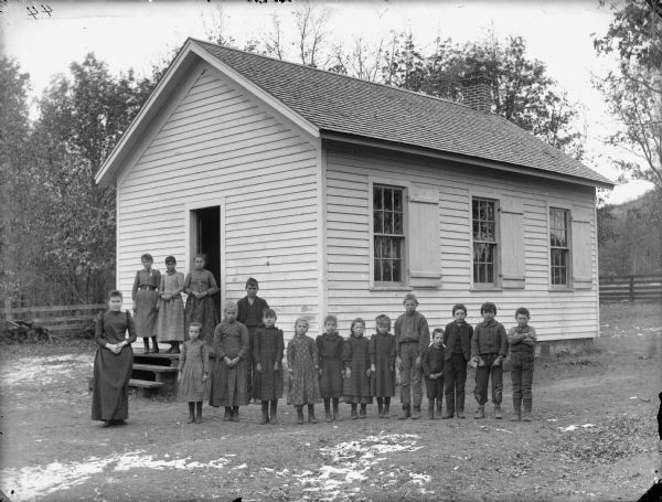 Group portrait of a woman and a group of boys and girls posing standing in front of a wooden building, probably a school group.	
