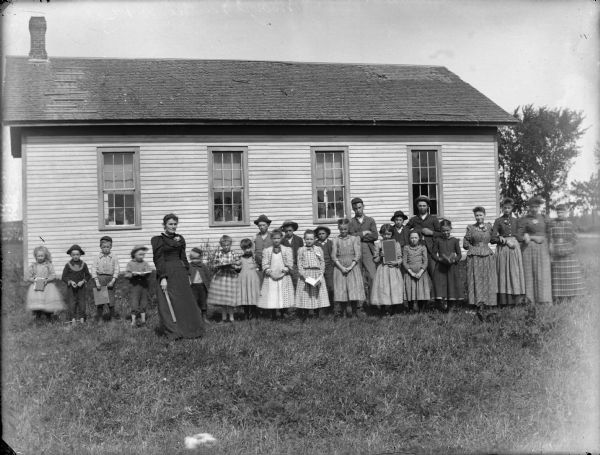 View of a woman holding a ruler standing with a group of boys and girls who are posing standing in front of a wooden building, probably a school group.