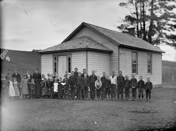 Group portrait of a woman and a group of boys and girls posing standing in front of a wooden building, probably a school group.	