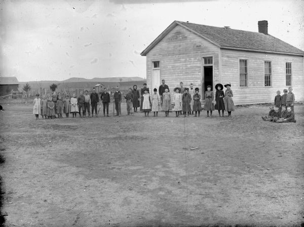 View towards a man and woman, and a group of boys and girls, posing standing in front of a wooden building, probably a school group.	