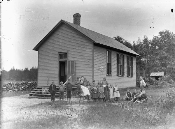 Group portrait of a woman and a group of boys and girls posing sitting and standing in front of a wooden building, probably a school group, possibly the Town Creek School.