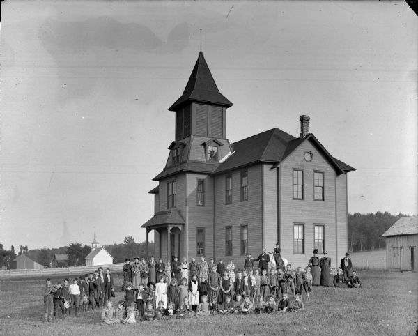 Group portrait of men, woman and primarily a group of boys and girls posing standing in front of a two-story wooden building, probably a school group and possibly the town school building.	