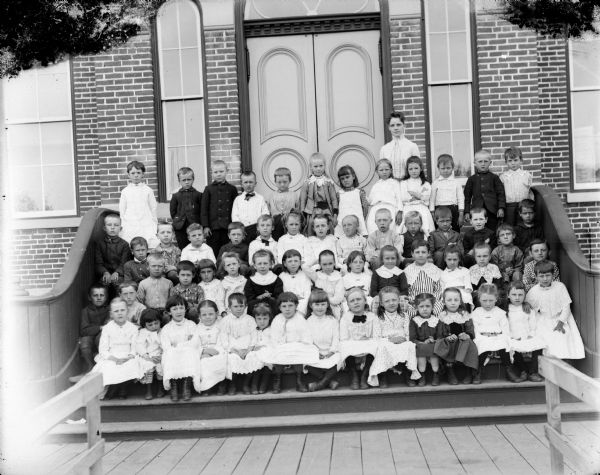 Woman and a group of boys and girls posing standing and sitting on the steps in front of a brick building. Probably a school group and possibly the Black Rivers Grade School, formerly the high school.