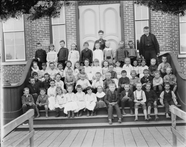 Group portrait of a woman and a group of boys and girls posing standing and sitting on the steps in front of a brick building, probably a school group and possibly the Black Rivers Grade School, formerly the high school. Identified, second from left on the top row, Oce Thompson (later Mrs. Frank Lozier), Class of 1898; fourth from left on the top row, Grace Jones (later Mrs. Emil Meck), Class of 1897; fourth from left on the second row, Peter K. Peterson; and seventh from the right on the second row, Carl Oyden.