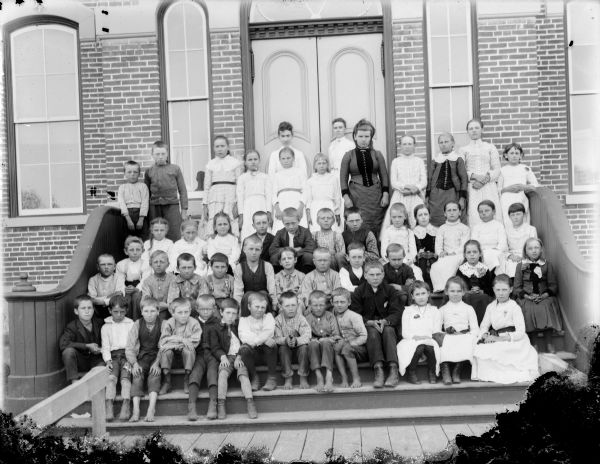 Two women and a group of boys and girls posing standing and sitting on the steps in front of a brick building, probably a school group and possibly the Black Rivers Grade School, formerly the high school.