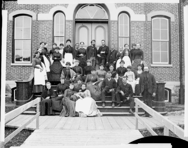 Group of men and women posed standing and sitting on the steps in front of a brick building, probably the Black Rivers Grade School, formerly the high school.	