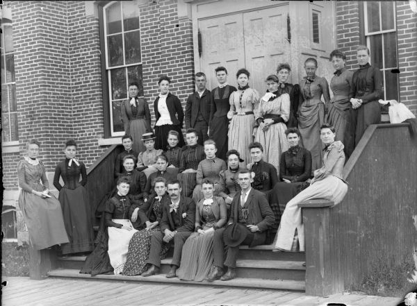 Group of men and women posed standing and sitting on the steps in front of a brick building, probably the Black Rivers Grade School, formerly the high school.