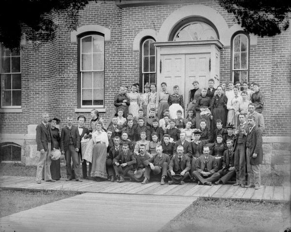 Group of men and women posed standing and sitting on the steps in front of a brick building, probably the Black Rivers Grade School, formerly the high school, possibly a meeting of the Teachers Institute. Identified in the group are Herbert Perry (seated, first row, third from the left with a moustache); Freeman Dell (seated, first row, sixth from the left with a moustache); and Edith Davis (third row and second from the right).