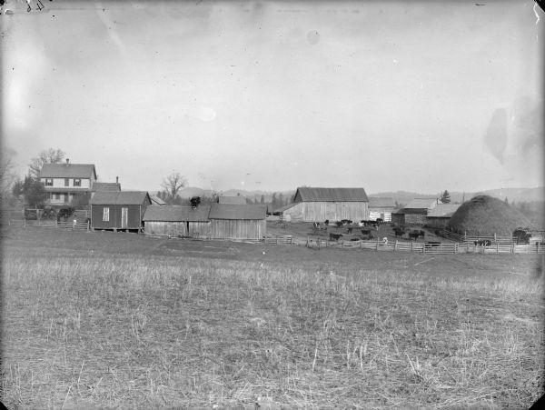 View across field of farm buildings, a frame house, cattle in a corral, and a large haystack. A group of people and buggies is posing near the farmhouse.	