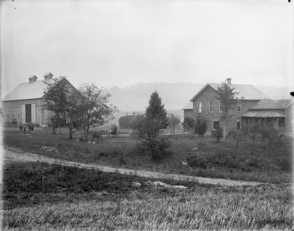 View across dirt road of farmyard. A man on the left stands with two horses in front of a barn. In the center there appears to be two women  sitting in a buggy pulled by a single horse, while two blurred horses are grazing near trees nearby. In the background a man and another persn are standing on a large haystack. On the right is a frame house.