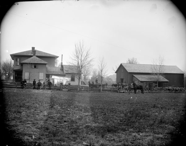 View across field towards men posing near a picket fence. They are standing near several wagons and horses near a two-story frame house and a barn.