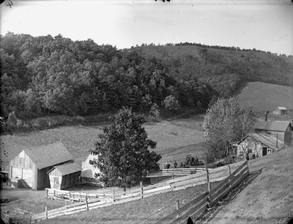 Elevated view from hill of farmhouse, farm buildings, and fenced yards. People are posing standing near the bottom of the hill behind a fence in the center, and another group of people is posing on the far right in front of the farmhouse.