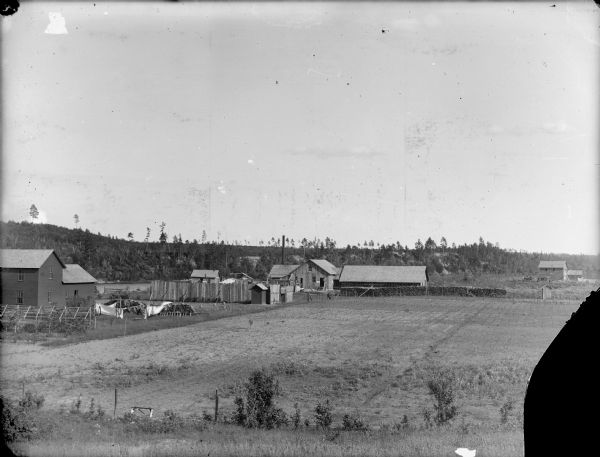 View across fields of a frame house and farm buildings, with a large supply of logs, near a forested area.