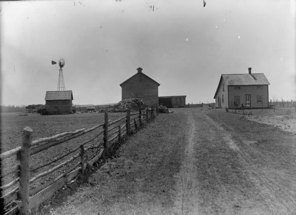 View along fence and road towards a frame house, barn, farm buildings, wooden fence, and a windmill.