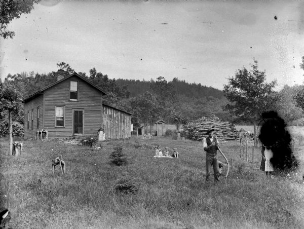 Man posing standing and holding/sharpening a scythe, with a dog sitting nearby. Three children are sitting in the grass, and a woman is standing in front of a frame house with another child by her side. On the far right an individual wearing a dress is obscured due to emulsion decay. In the background near a large pile of wood is a horse and buggy.