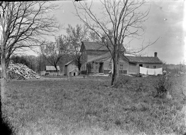 View across grass towards a board frame house, farm buildings, hanging laundry, and a large wood pile.	