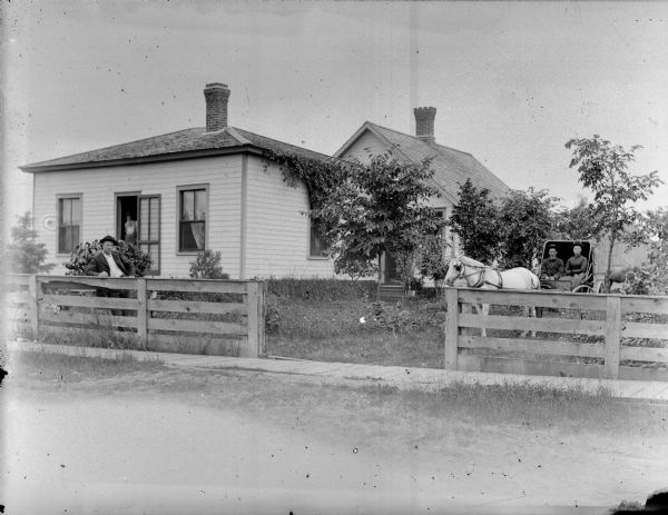 Man posing standing near a wooden fence surrounding a frame house. Another man is posing standing in the doorway, while a man and woman are posing sitting in a buggy pulled by a horse.