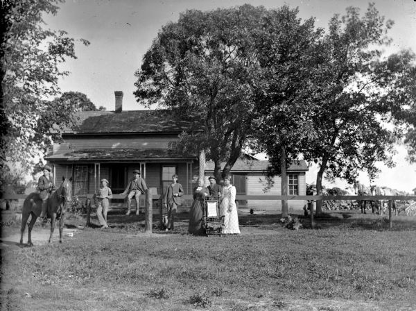 Two women, two men, and three boys posing near a wooden fence in front of a frame house. One boy is sitting on the fence and another is mounted bareback on a horse.