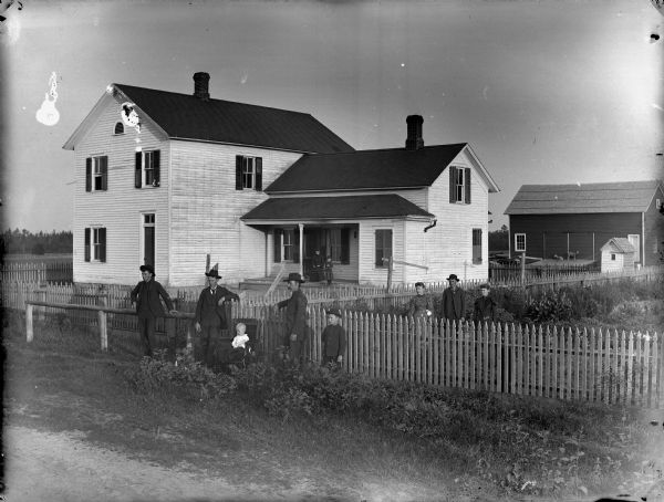 Four men, two women, a boy, and a small child in chair are posing near a picket fence in front of a frame house, with a woman and girl posing on the porch.