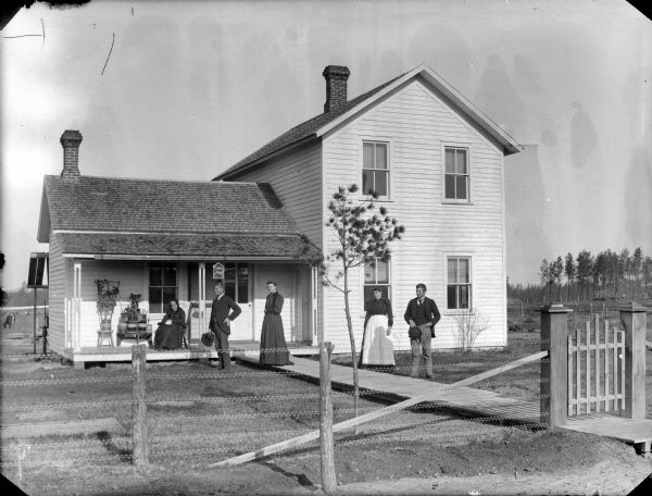 Two men and two women posing standing in the yard of a frame house. Behind them an elderly woman is sitting on the porch.